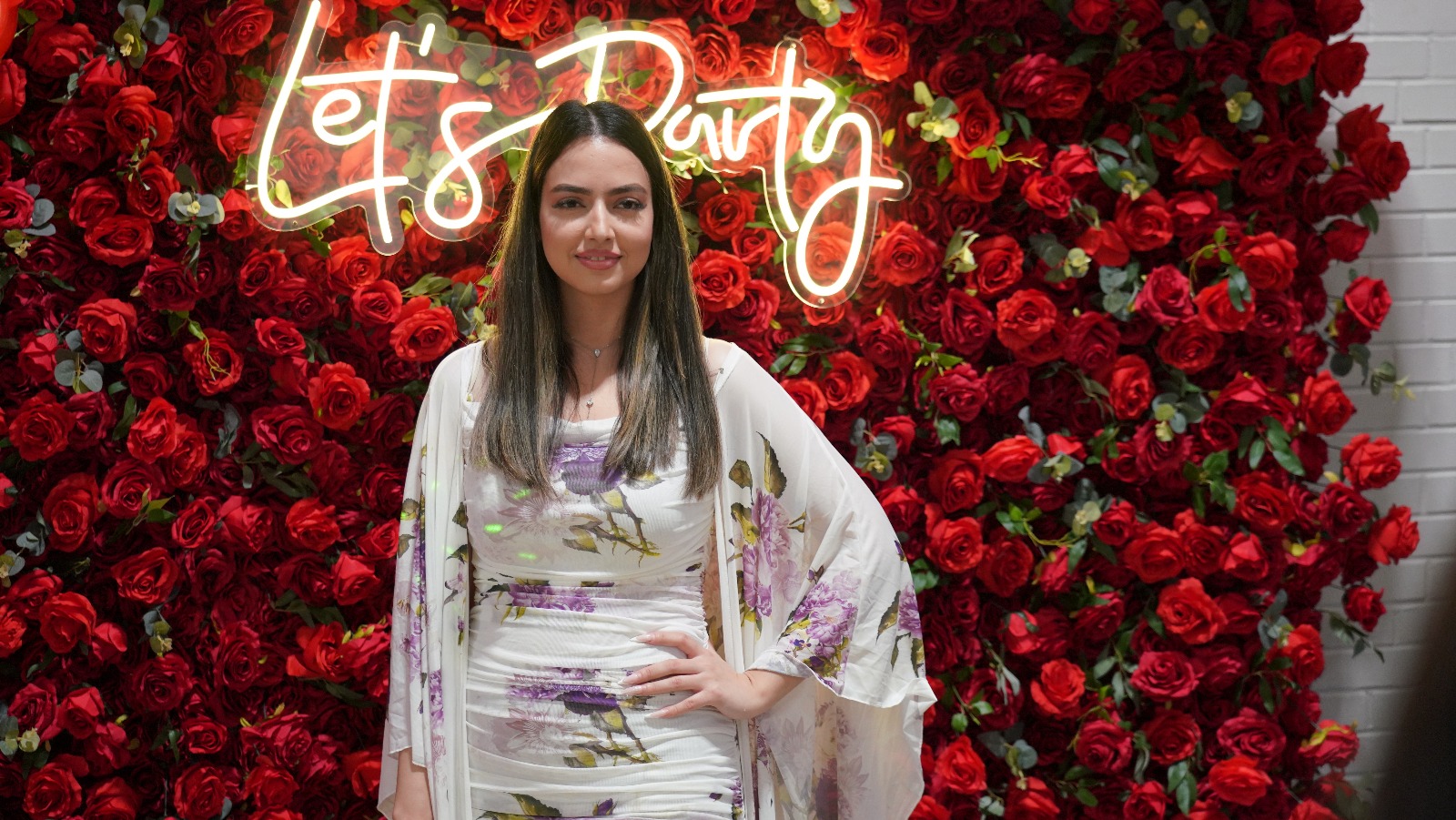 Woman in a floral dress before a red rose wall with a neon Let's Party sign