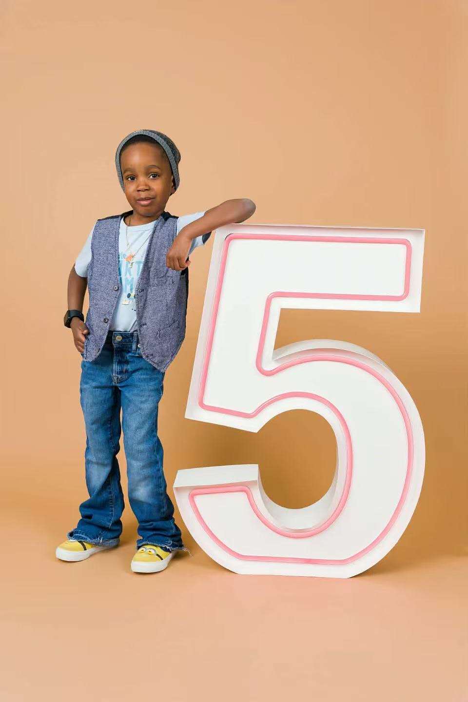 Young boy leaning on a giant marquee number 5 with pink neon trim