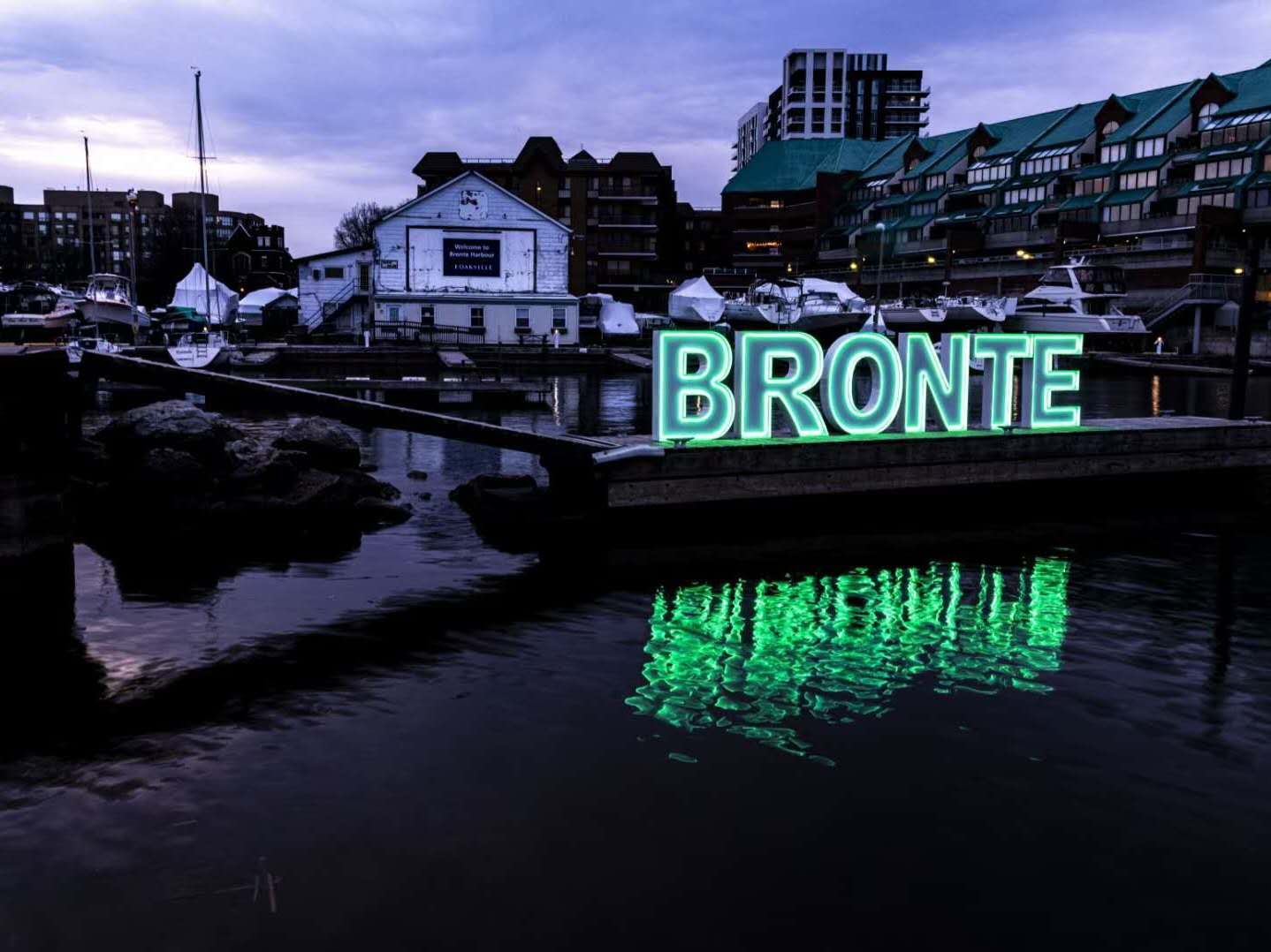 Green neon BRONTE letters on a harbour dock at dusk with water reflection