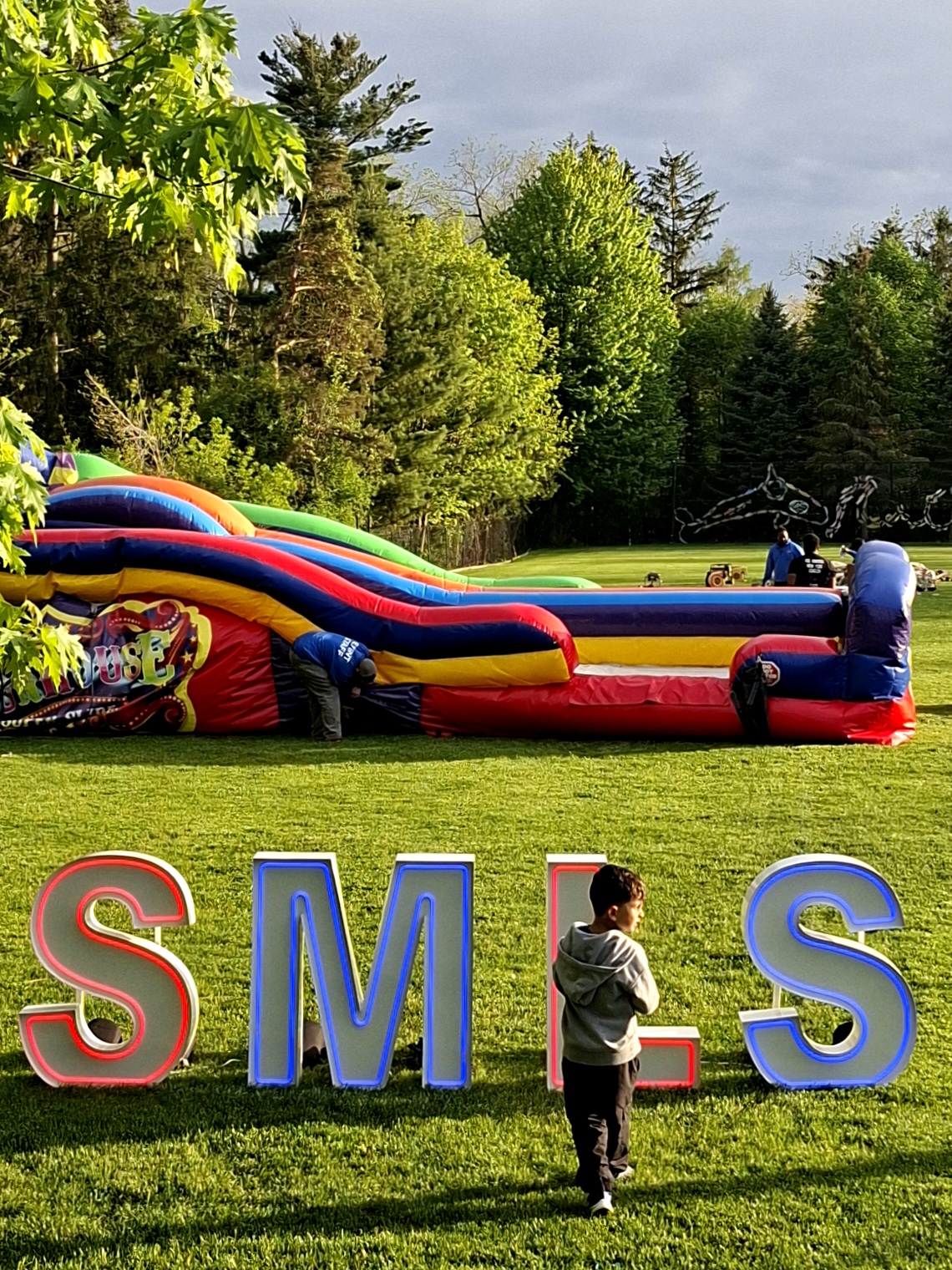 Child beside giant neon SMLS letters with an inflatable slide on a lawn