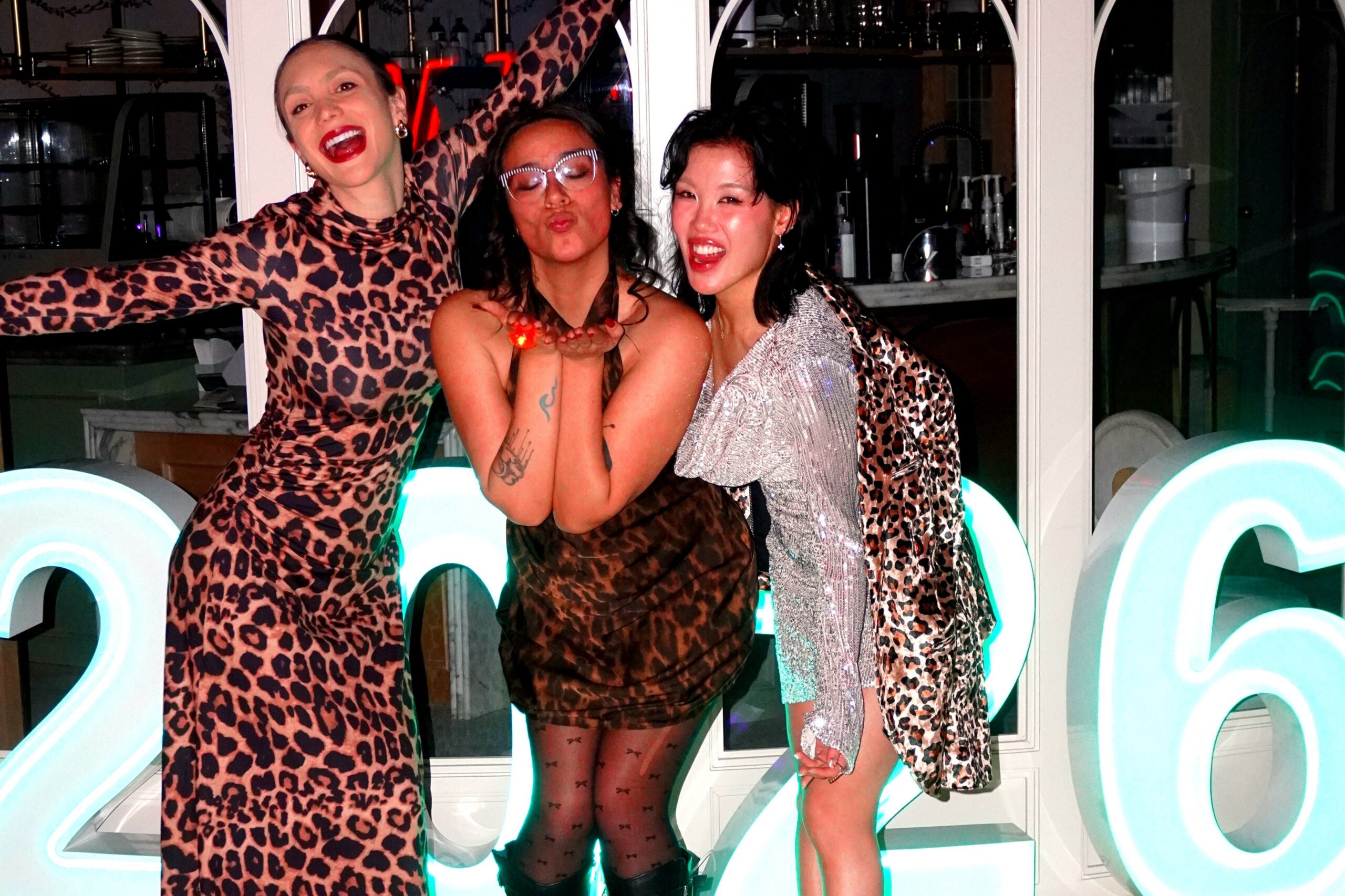 Three women posing playfully before illuminated 2026 numbers at a party