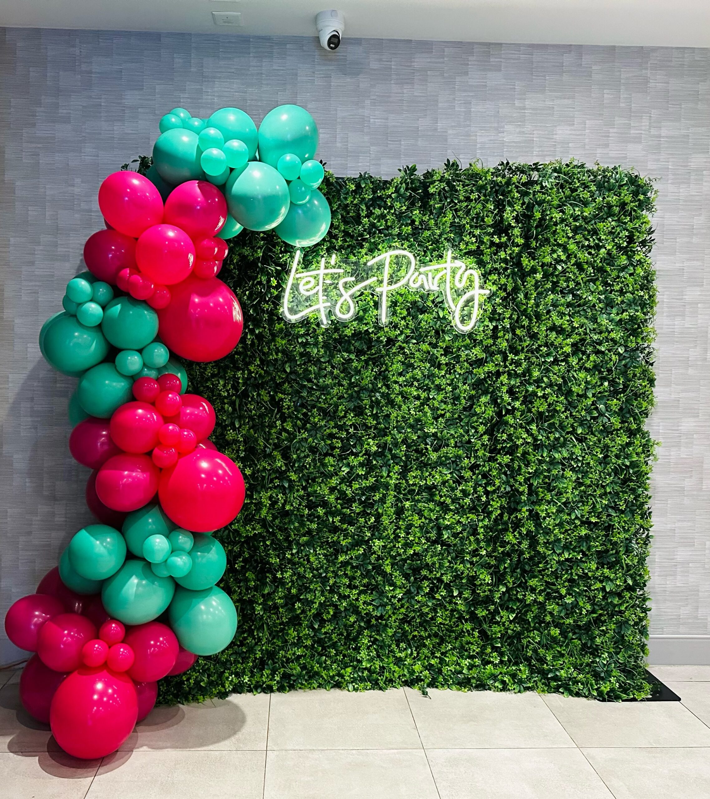 Green hedge wall with a Let's Party neon sign and balloon garland