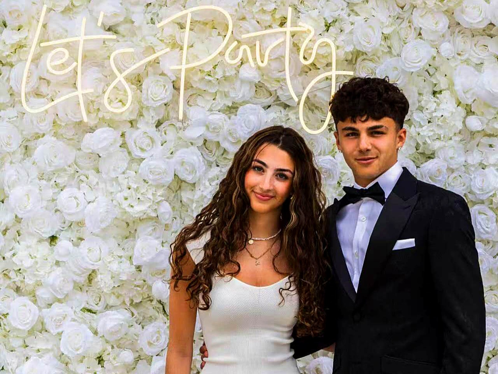 Couple at white flower wall with Let's Party neon sign